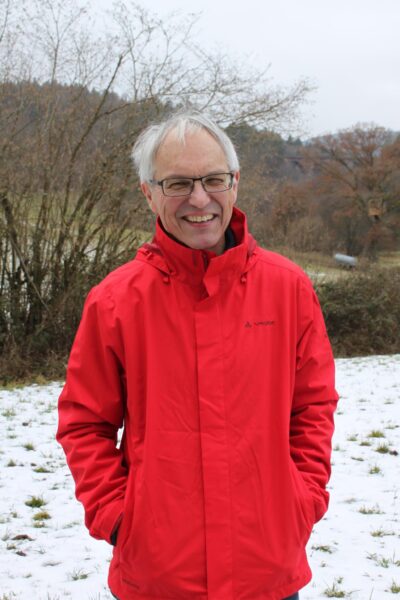 Horst Kowarsch steht in einem roten Anorak auf einer verschneiten Wiese und lacht in die Kamera.